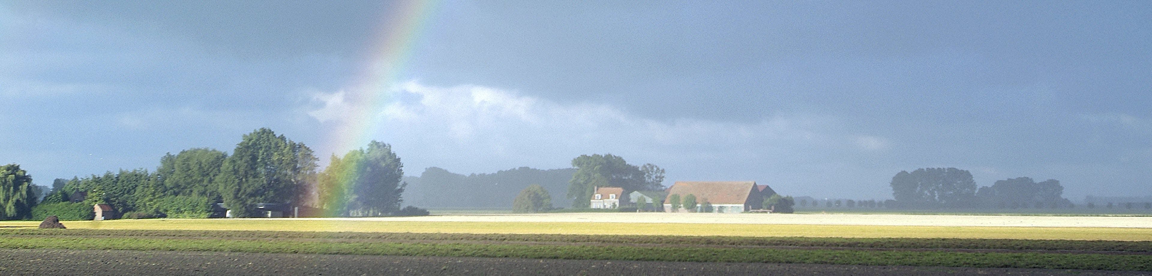 Een boerderij nabij een akker. In de lucht is een regenboog te zien.