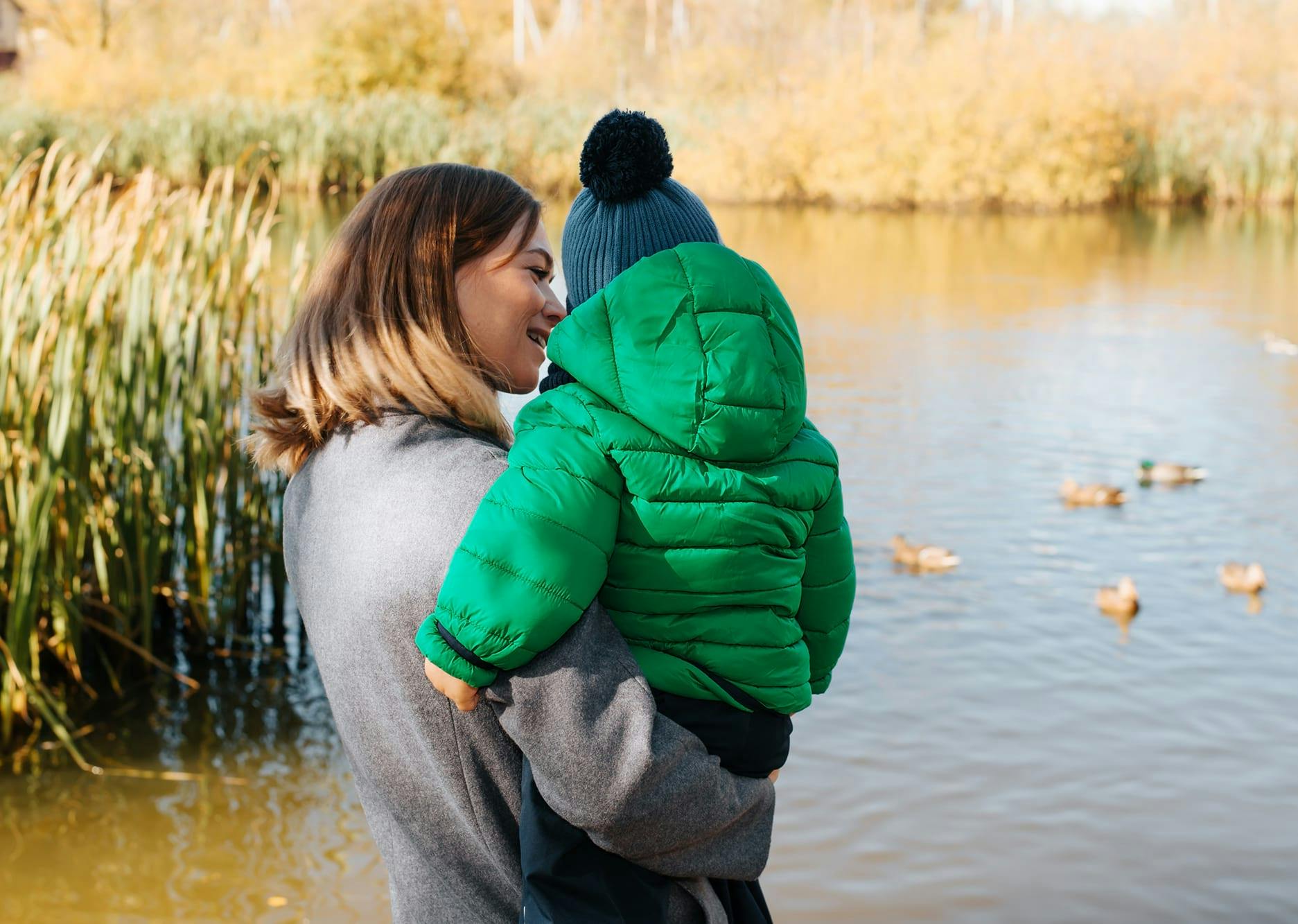 Vrouw staat met een kindje op de arm te kijken naar eendjes die in het water zwemmen