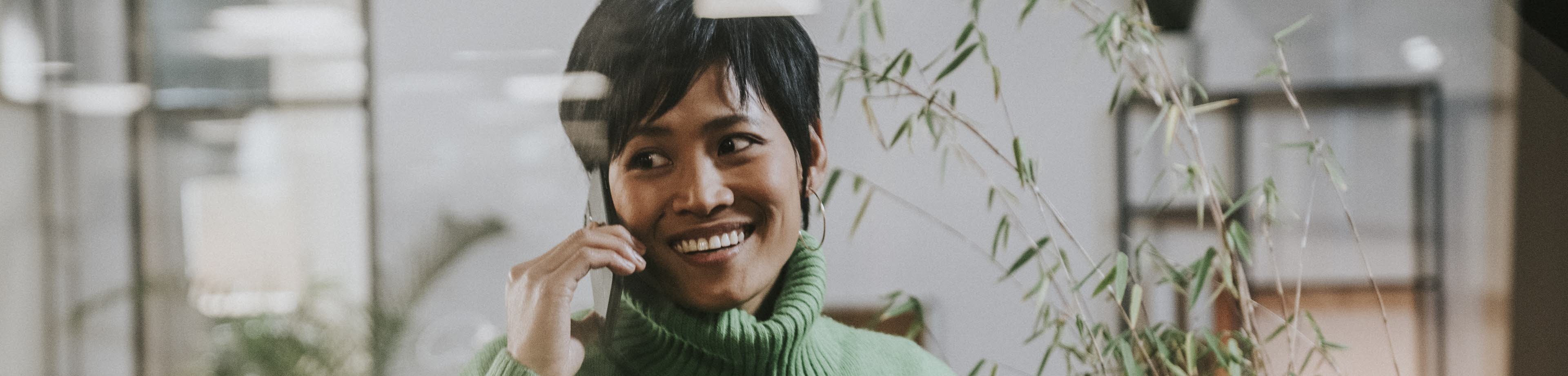 Een vrouw met een groene trui glimlacht en houdt een mobiele telefoon tegen haar oor.