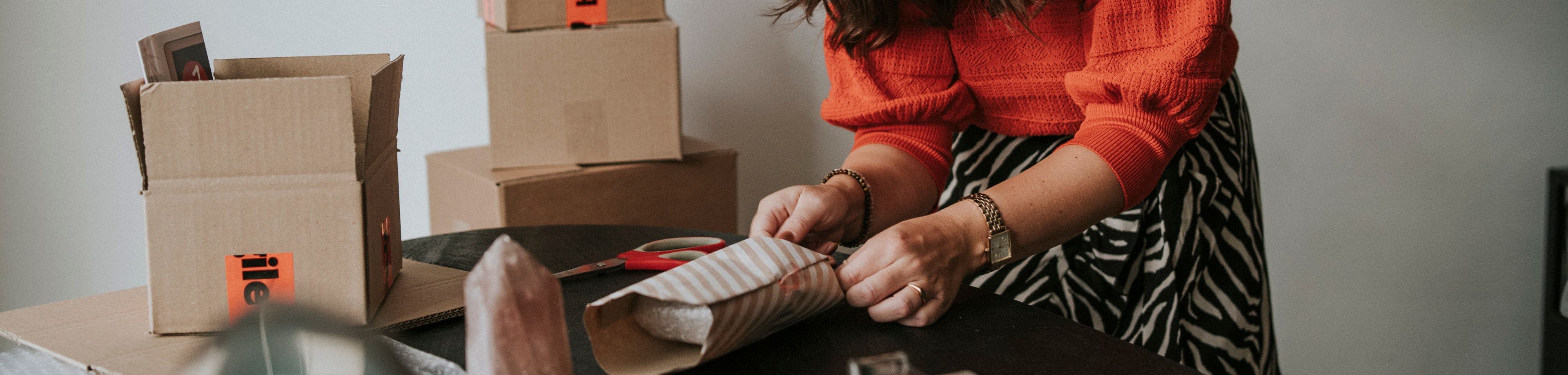 Een vrouw met een rood-oranje trui maakt op een tafel een pakketje verzendklaar. Er staat een aantal verzenddozen op en naast de tafel.