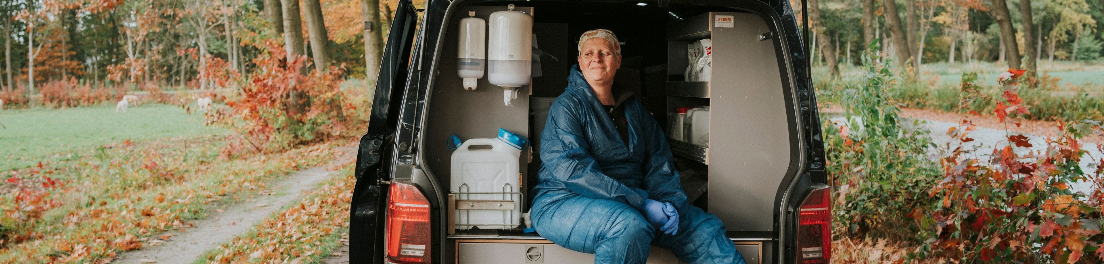 Dierenarts Janny Hermans zit ontspannen op de achterdorpel van een bedrijfswagen, die in een bosrijke omgeving geparkeerd staat.