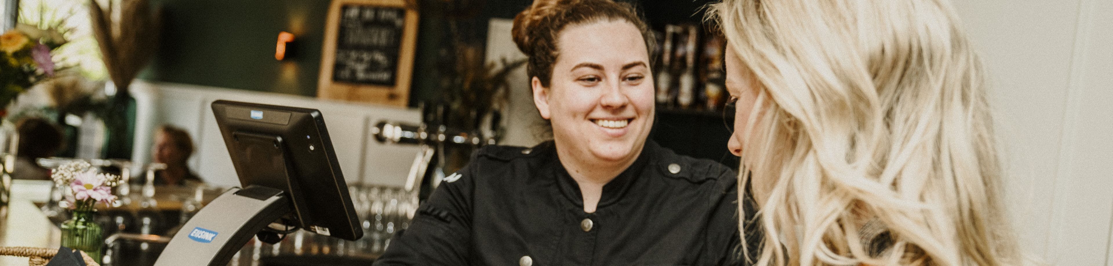 Een klant van een horeca-gelegenheid rekent met haar pinpas af bij een vrolijk lachende medewerker.