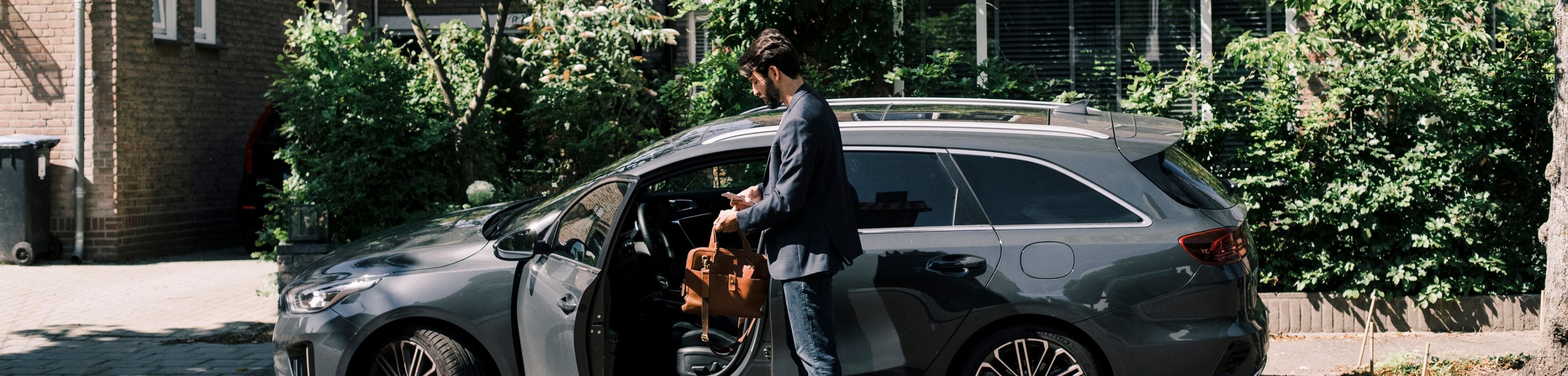 Een man staat naast zijn auto met geopend portier. Hij heeft zijn tas en telefoon in de hand, en is klaar om in te stappen.