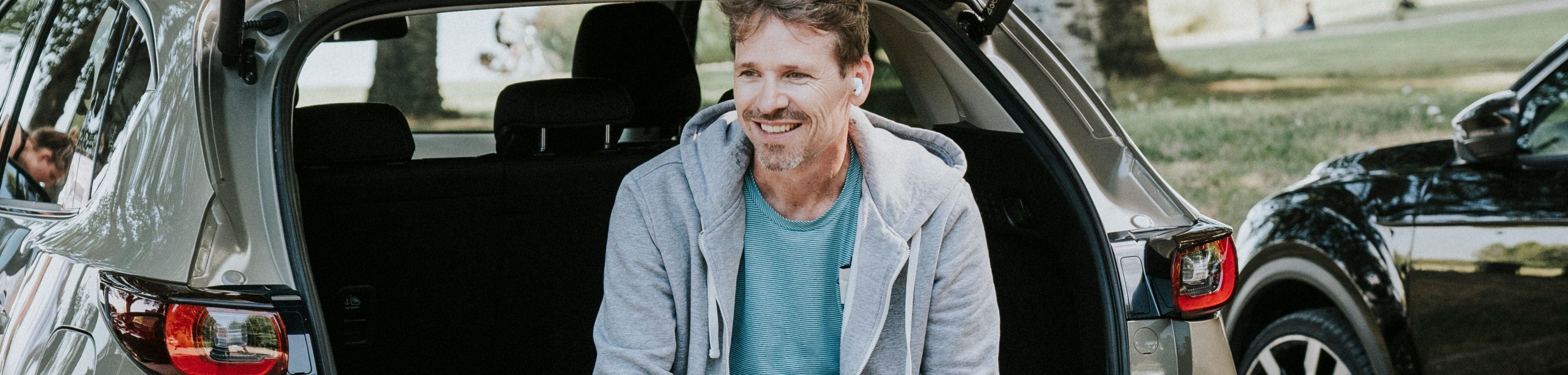 Man zit achter op de rand van de auto met de achterklep open en heeft een hond vast