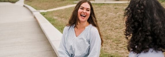 Een vrouw zit op de betonnen rand van een perk en lacht uitbundig naar een andere vrouw, met wie ze in gesprek is.