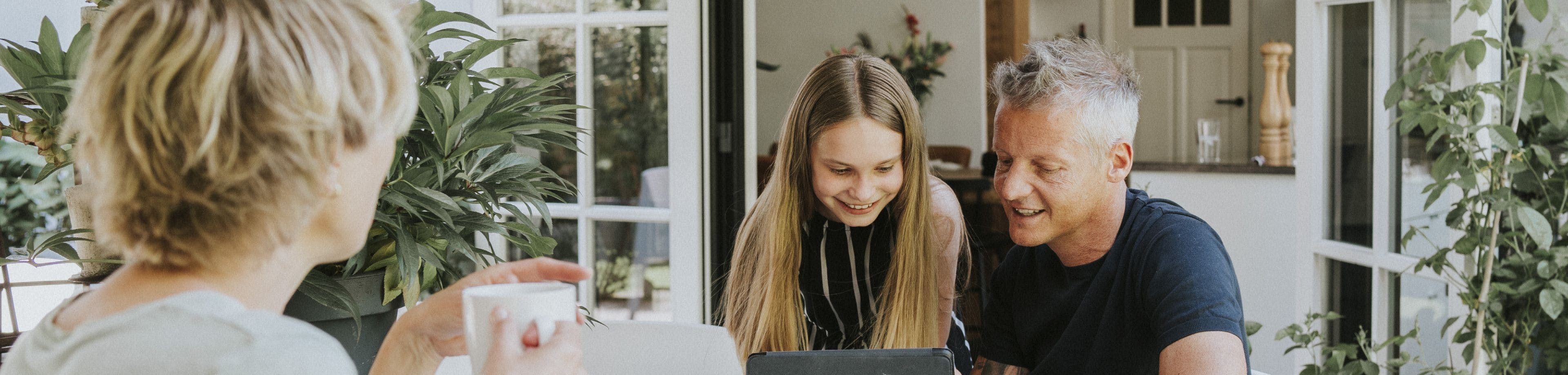 Vader zit samen met dochter met een laptop aan tafel. Moeder zit erbij met een kop thee.