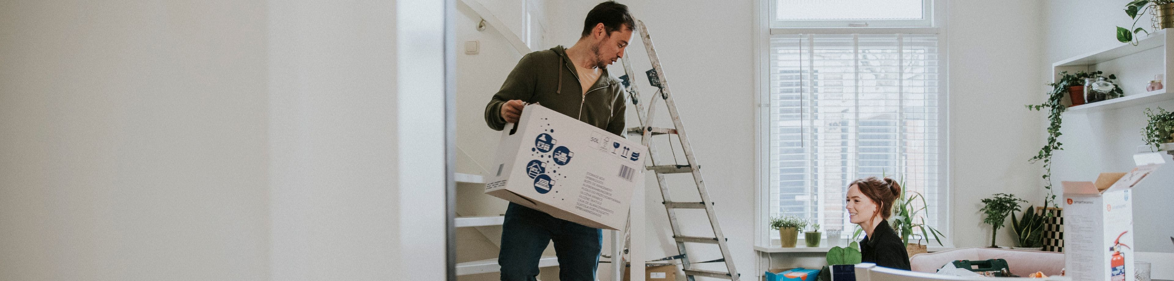 Vrouw zit op de vloer naast een trap. Man loopt met een verhuisdoos in zijn handen.