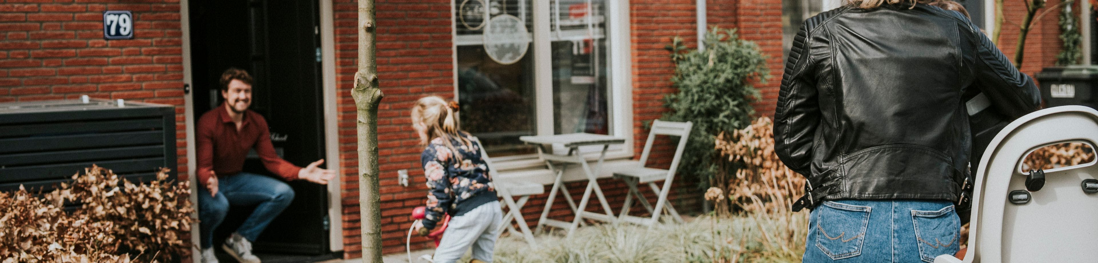 Een kind fietst van een vrouw op de fiets naar een man die op zijn hurken in de deuropening zit