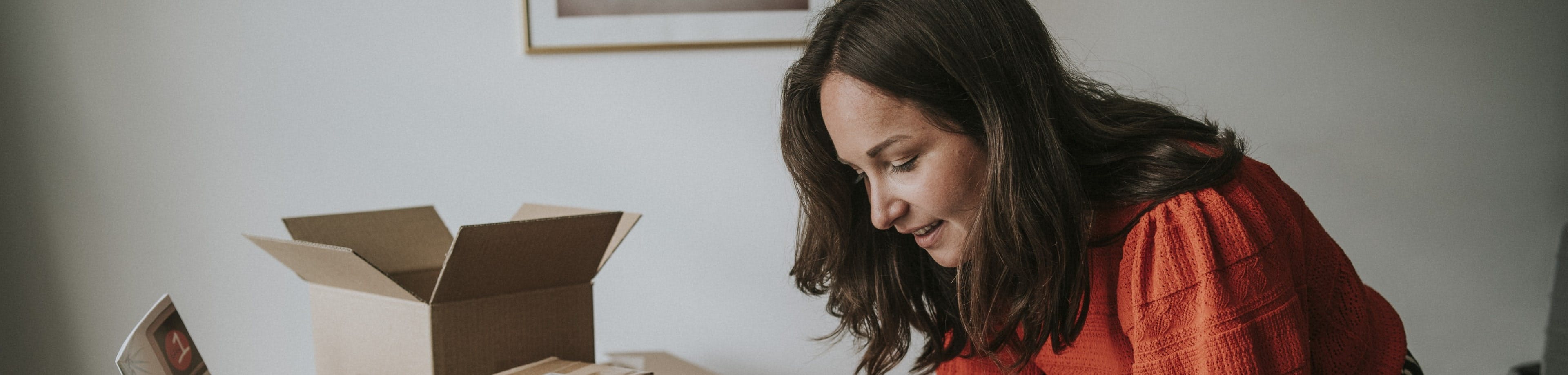 Een glimlachende vrouw met een rood-oranje trui buigt zich in een thuiswerkomgeving over haar werkplek. Ze schrijft iets op een papieren schrijfblok. Naast haar op het bureau staan enkele kartonnen dozen.