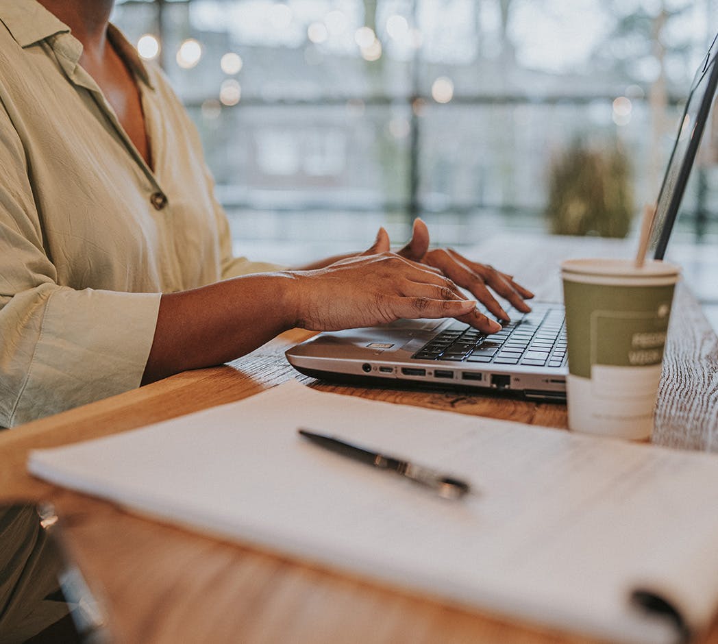 Iemand typt op het toetsenbord van een laptop. Naast de laptop staat een koffiebeker, en op de voorgrond ligt een schrijfblok met een pen.