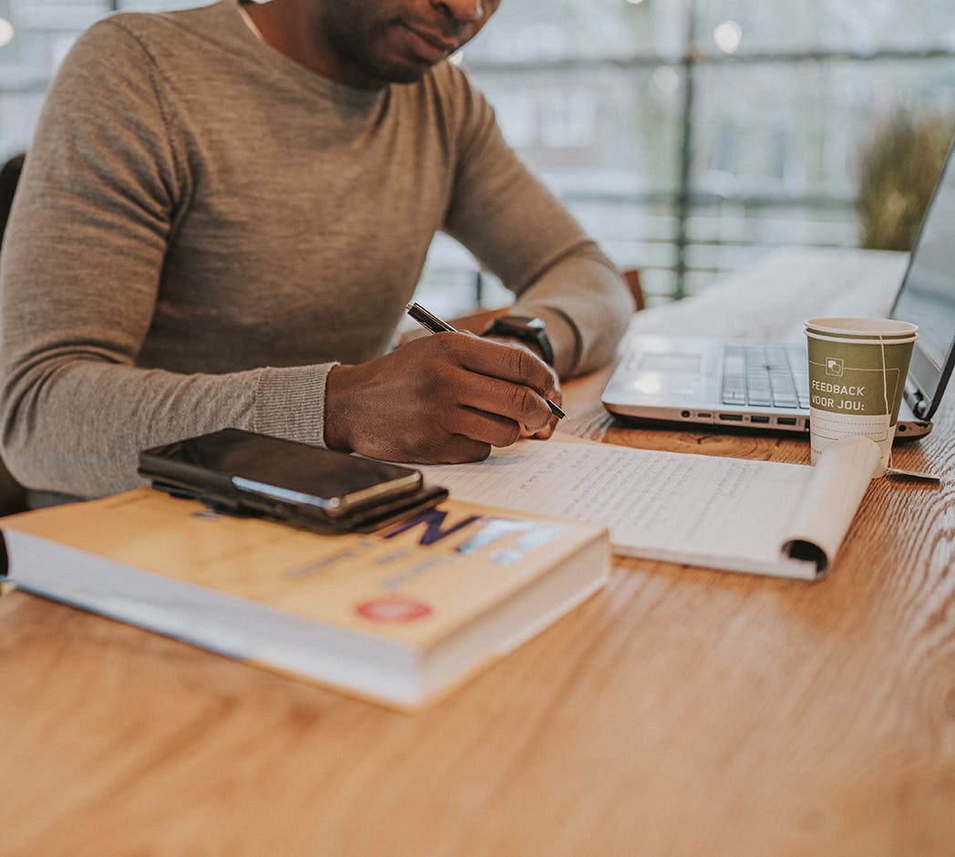 Iemand zit aan een tafel met daarop een openstaande laptop. Deze persoon schrijft op een schrijfblok dat naast de laptop ligt.