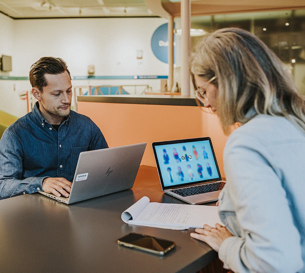 2 mensen zitten tegenover elkaar en hebben een laptop open staan. 1 van hen kijkt op het laptopscherm, de ander leest de tekst die op een notitieblok geschreven staat.