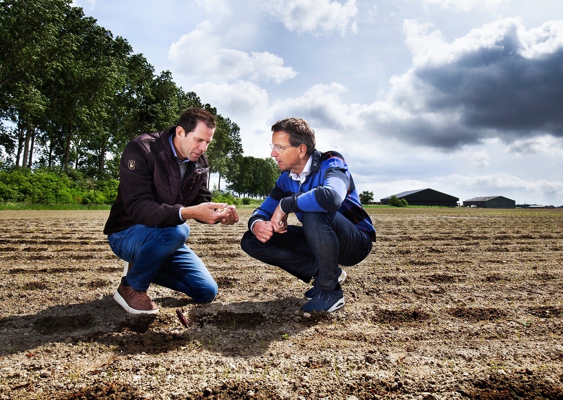 2 mannen zitten gehurkt op een akker en zijn in gesprek met elkaar.
