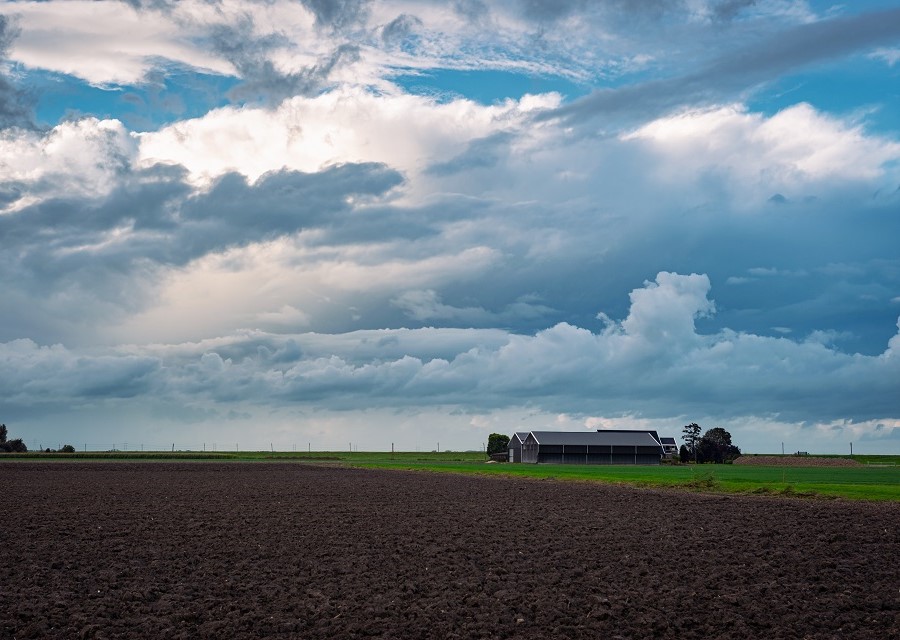 Een braakliggende akker met enige bebouwing, onder een stormachtige wolkenlucht.