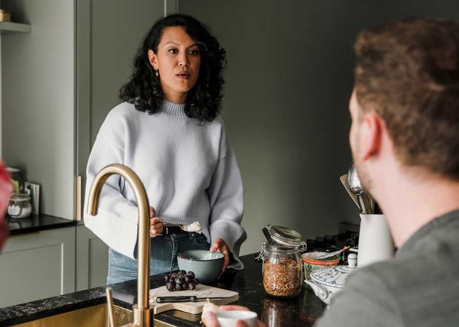 Een vrouw staat aan het aanrecht in de keuken, en is gesprek met een andere persoon.