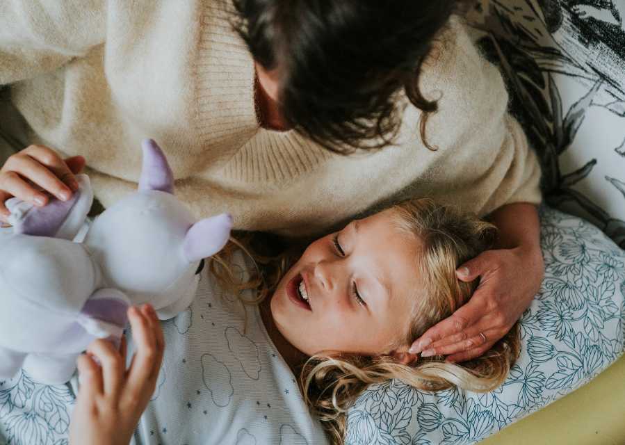 Moeder stopt haar dochter in bed in, dochter houdt een eenhoornknuffel vast.