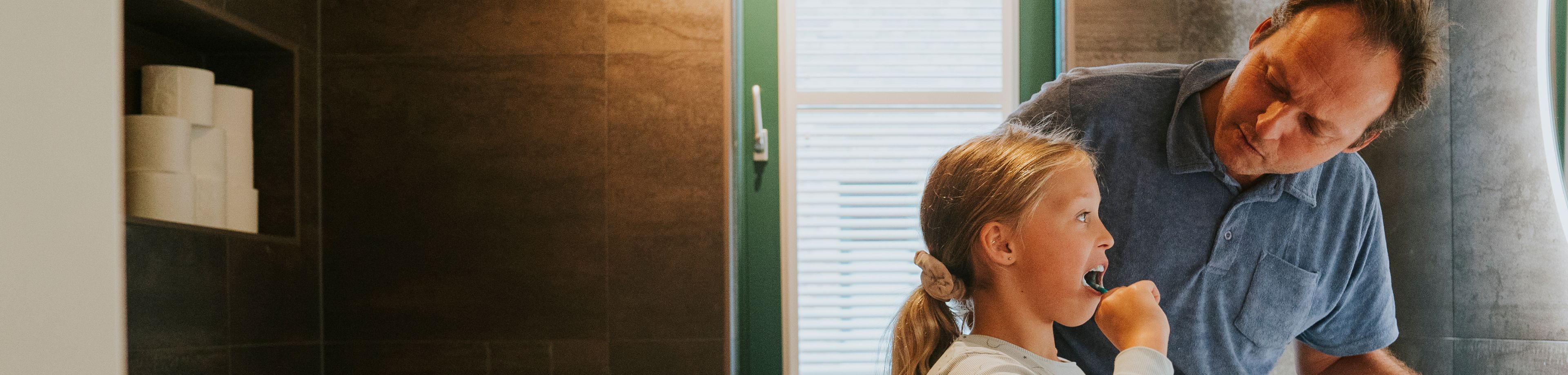 Meisje poetst haar tanden in de badkamer terwijl haar vader toekijkt. 