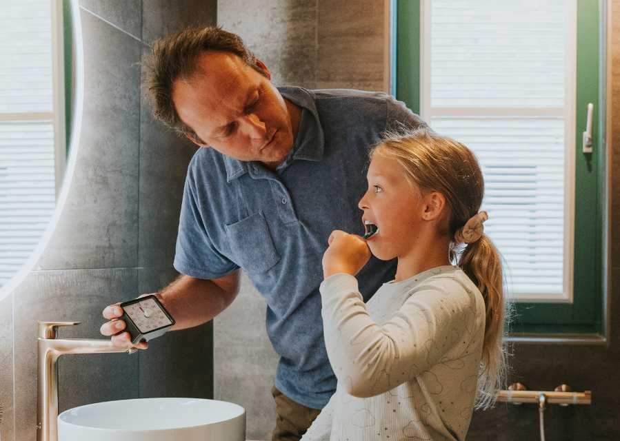 Vader houdt een timer bij terwijl dochter voor de spiegel in de badkamer haar tanden poetst.