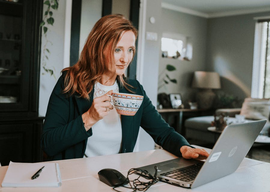 Een vrouw zit aan aan tafel met een grote kop thee in haar handen. Ze kijkt op de laptop die voor haar op de tafel staat.
