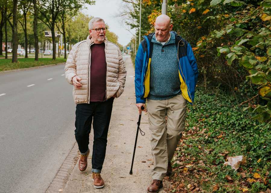 Twee mannen, waarvan een ondersteund door een wandelstok, lopen samen over de stoep.