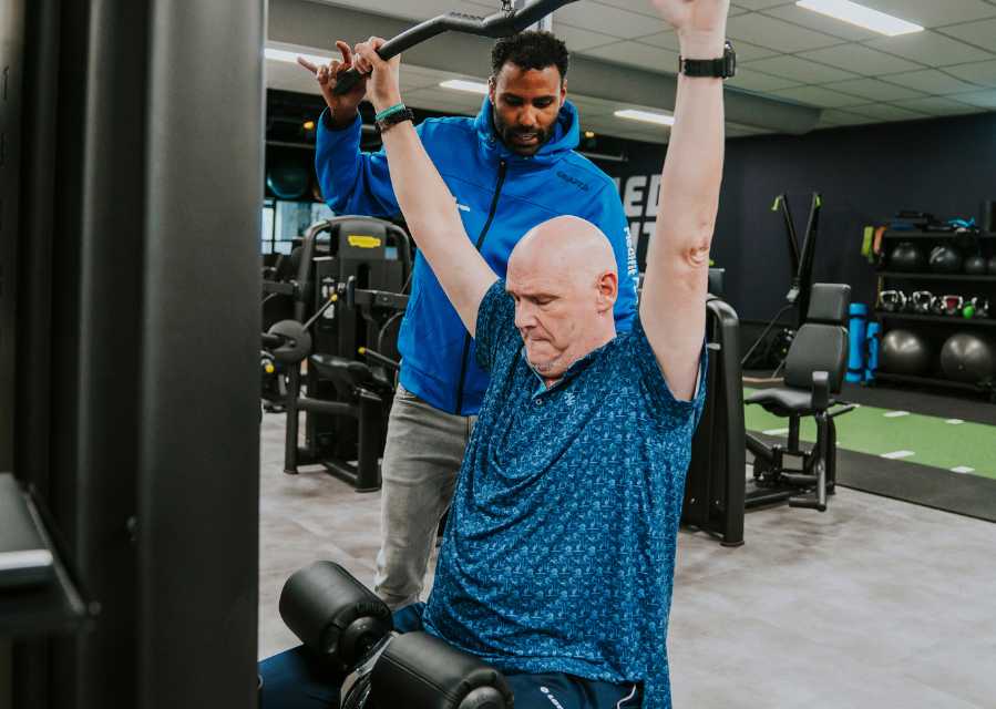 Een fysiotherapeut begeleidt een man met een krachtoefening op een fitnesstoestel. 