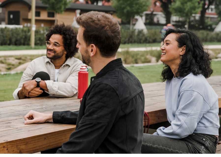Drie mensen zitten aan een houten tafel in een park.