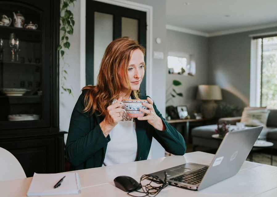 Een vrouw heeft een kop thee in haar handen, terwijl ze op de laptop kijkt die voor haar op tafel staat.
