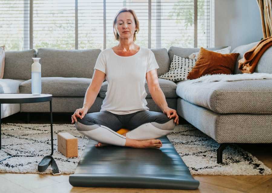 Een vrouw zit in een yogapositie te mediteren op een matje in de woonkamer.