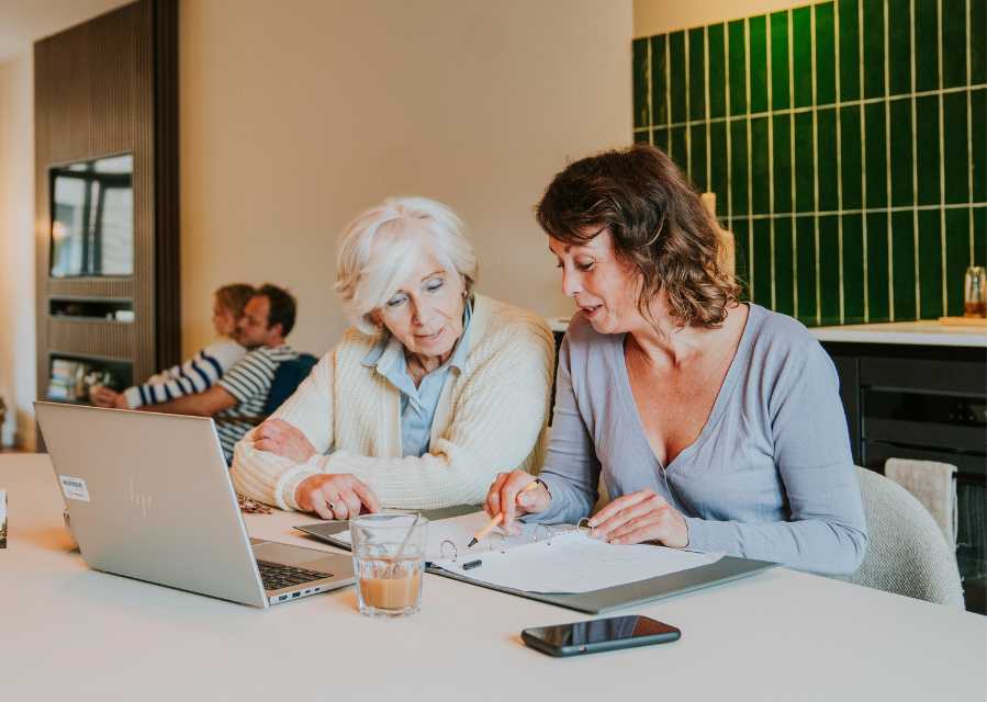 Moeder en oma zitten achter een laptop en bekijken een map met papieren documenten.