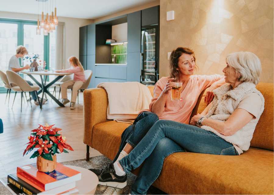 Moeder en oma zitten op een bank en dochter en zoon zitten op de achtergrond aan de keukentafel. 