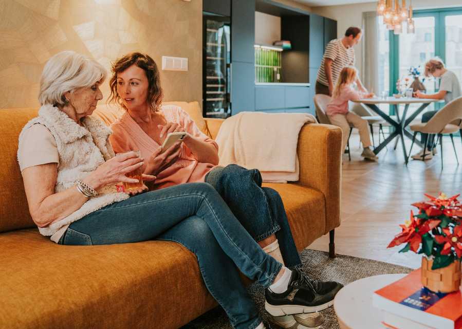 Moeder en oma bekijken een telefoon op de bank en vader en kinderen zitten aan de eettafel in de achtergrond.