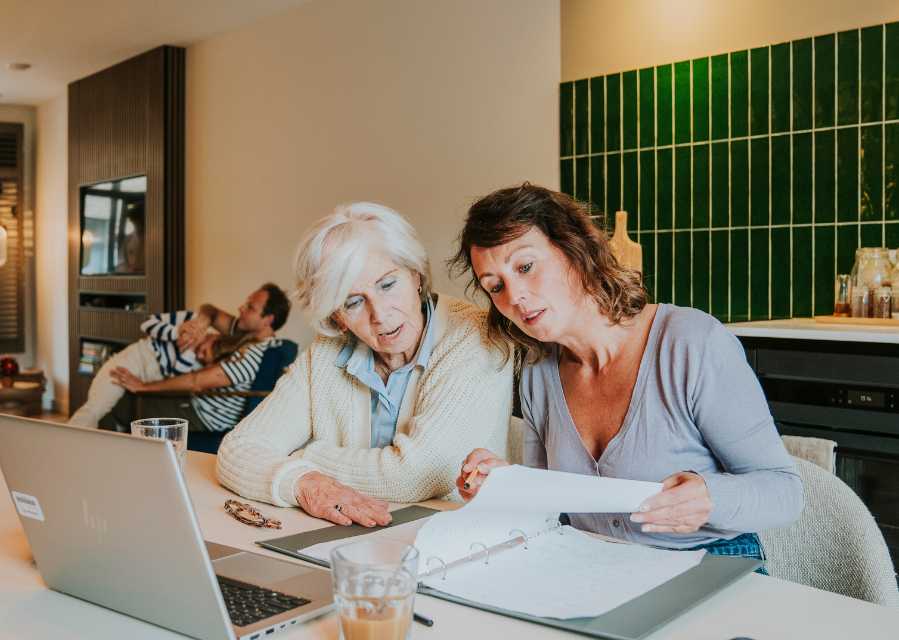 Moeder en oma bekijken samen een map met documenten aan de eettafel. 