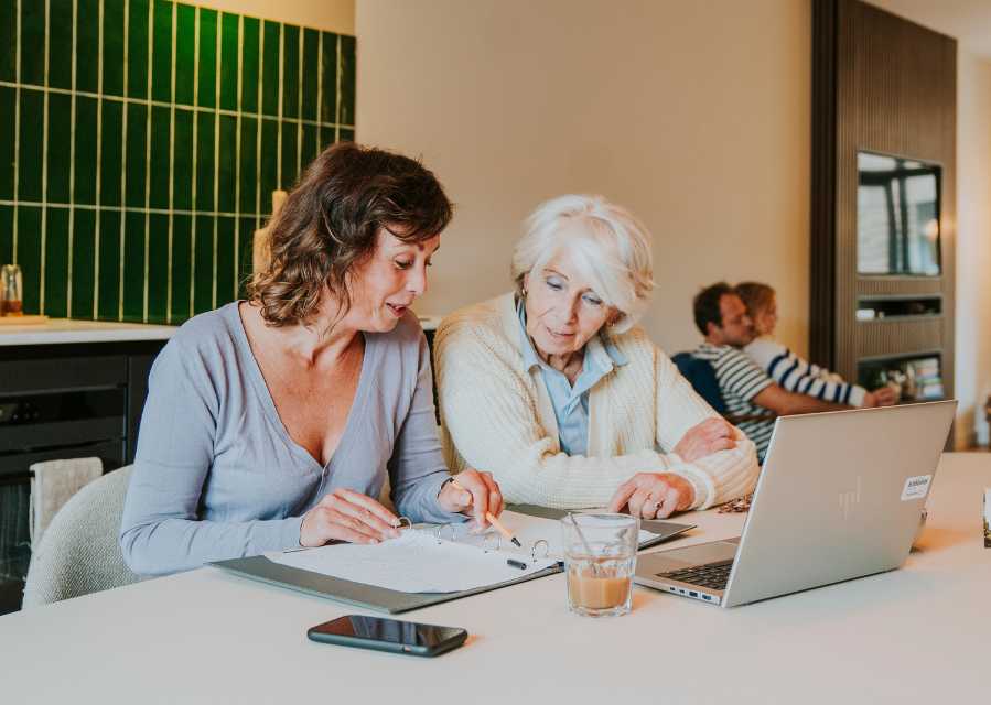 Moeder en oma zitten aan de eettafel achter een laptop en bladeren door een map met papieren documenten. 