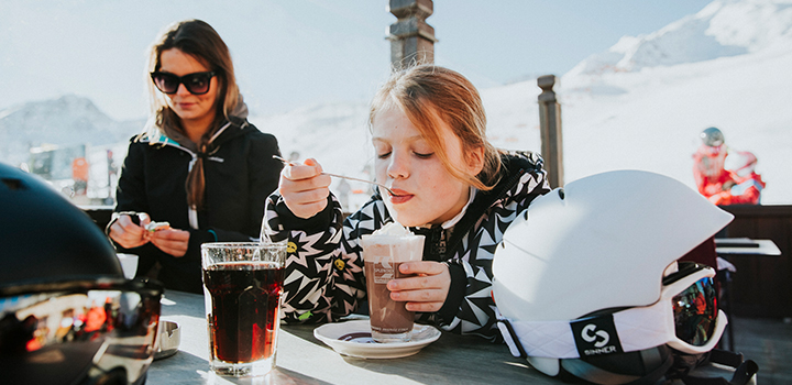 Kind op een terras op een skipiste met een warme chocomel met slagroom