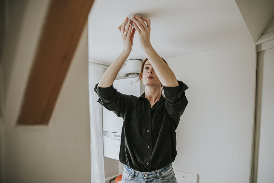 Een vrouw maakt een rookmelder aan het plafond schoon, zodat deze goed blijft werken.