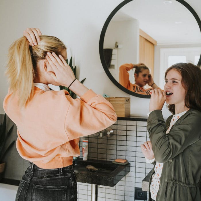 Twee jonge vrouwen staan bij een spiegel in de badkamer, de ene poets haar tanden, da ander doet haar haar