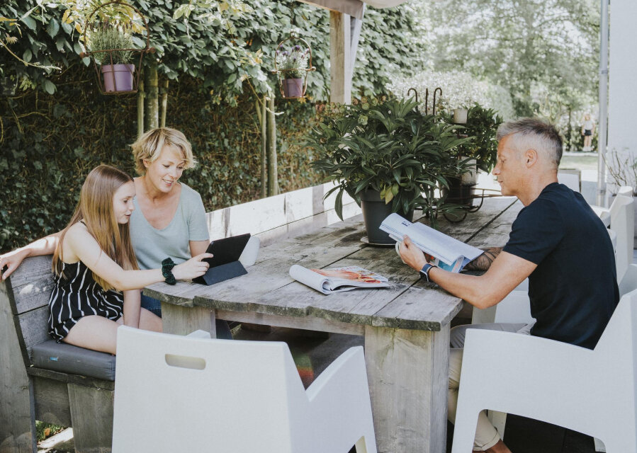 Een man, vrouw en een kind zitten aan een houten tuintafel. De man leest een tijdschrift, de vrouw en het kind kijken op een tablet.