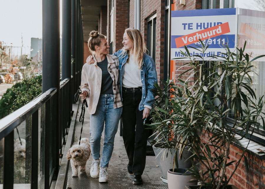 Twee vrouwen lopen schouder aan schouder over het balkon van een appartementengebouw. Ze kijken elkaar aan, en met 1 van hen loopt een hondje mee aan de riem.