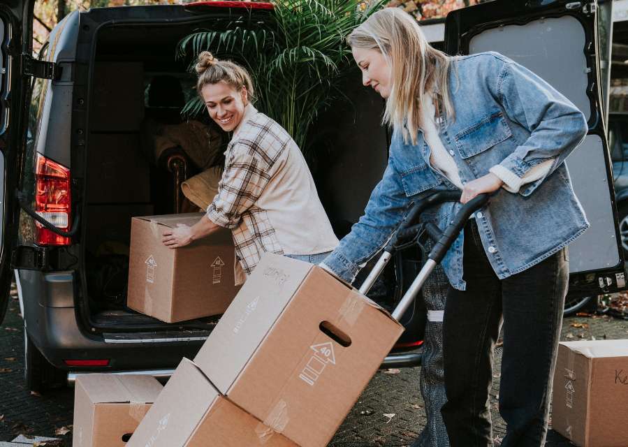 Twee vrouwen laden samen verhuisdozen uit een bestelauto op een steekwagentje.