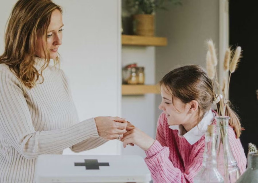 Een vrouw met een witte trui plakt een pleister op de vinger van een meisje. Op de voorgrond staat een witte ehbo-doos.