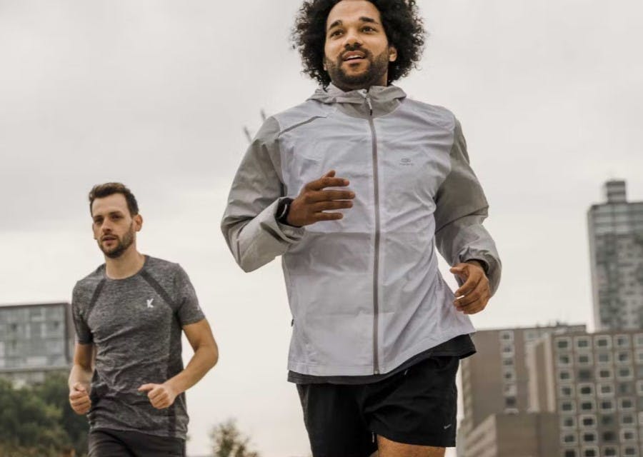 2 mannen in sportieve kleding zijn aan het hardlopen door de stad. Op de achtergrond zijn enkele hoge stadsgebouwen te zien.
