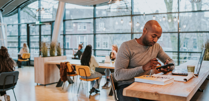Man typt op zijn telefoon terwijl hij in een kantoortuin achter zijn laptop zit