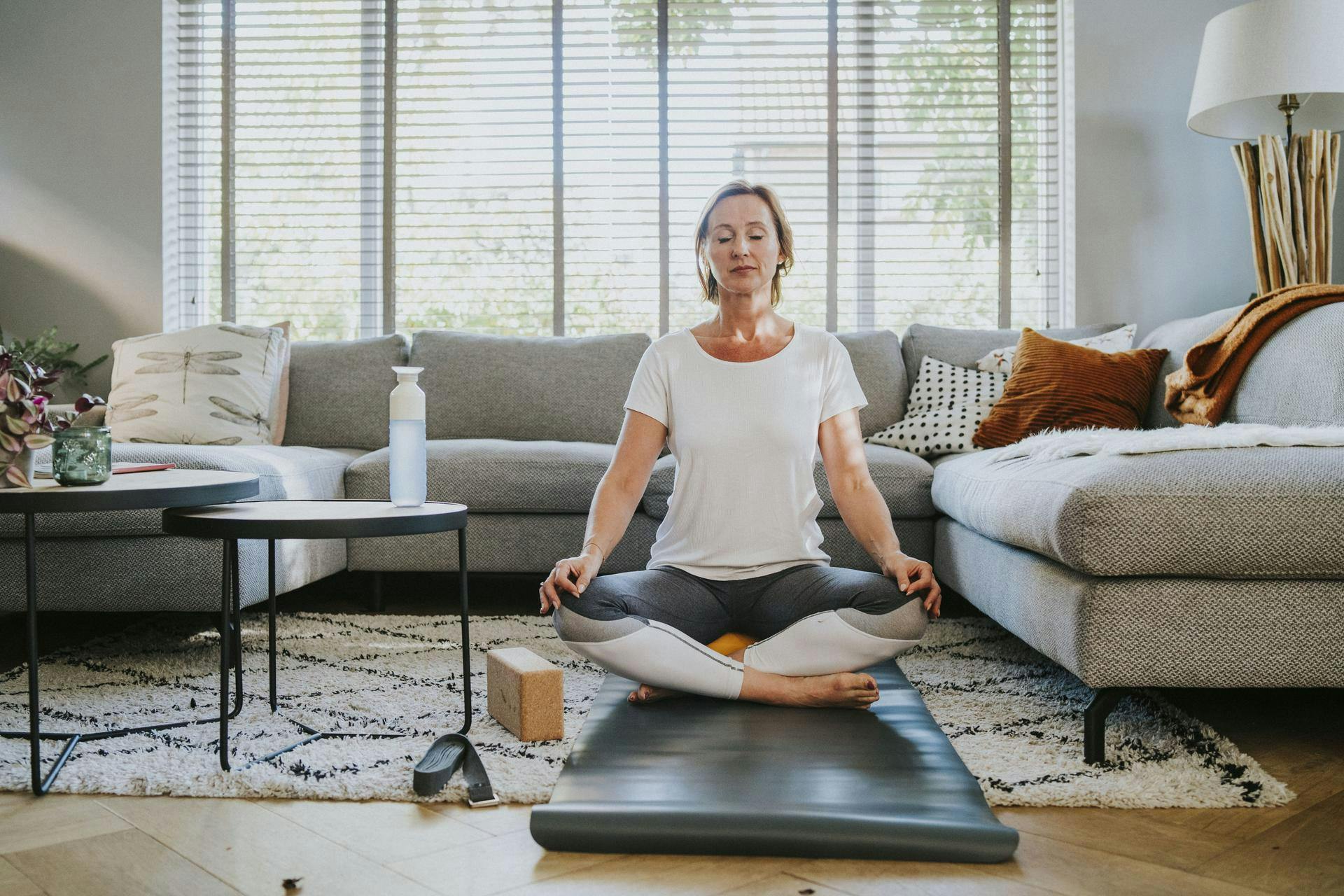 Een vrouw met een wit t-shirt zit op een yogamat in de woonkamer en mediteert met haar ogen dicht.