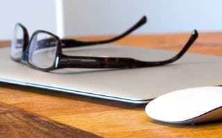Een bril, laptop en muis die op een houten tafel liggen.