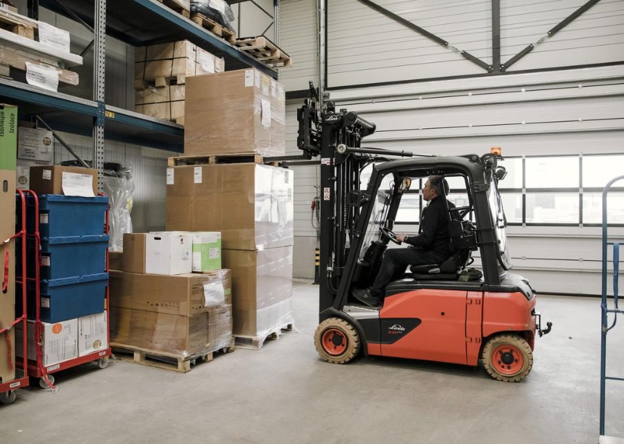 Een persoon bestuurt een vorkheftruck in een magazijn, en tilt daarmee een pallet met producten bovenop een ander pallet.