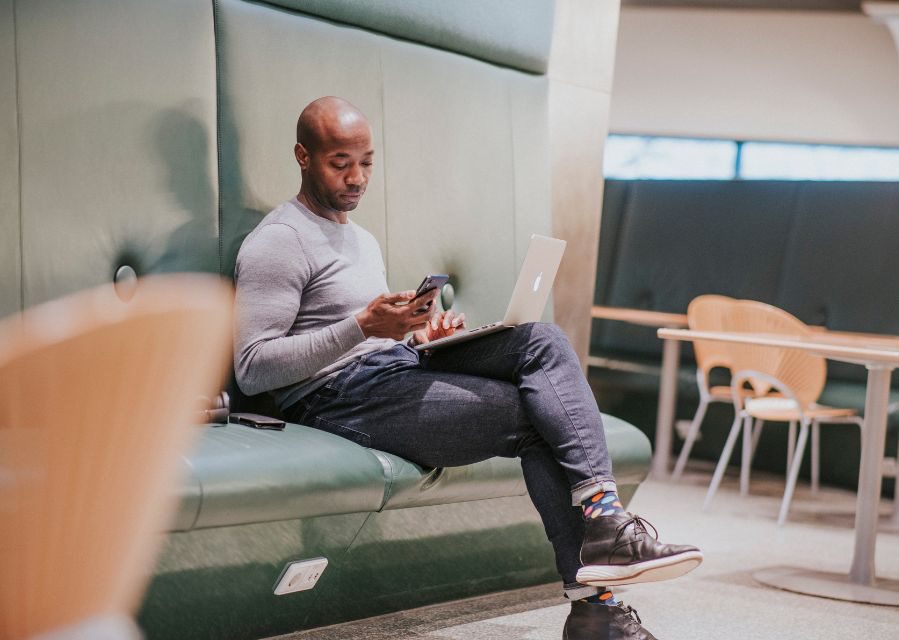 Een man zit op een bankje te werken; hij heeft een laptop op schoot en kijkt op de mobiele telefoon die hij in zijn hand heeft.