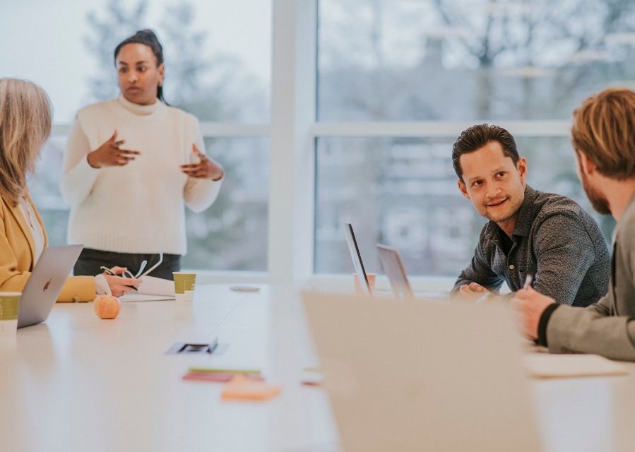 Vier mensen zitten of staan bij een vergadertafel en zijn met elkaar in gesprek.