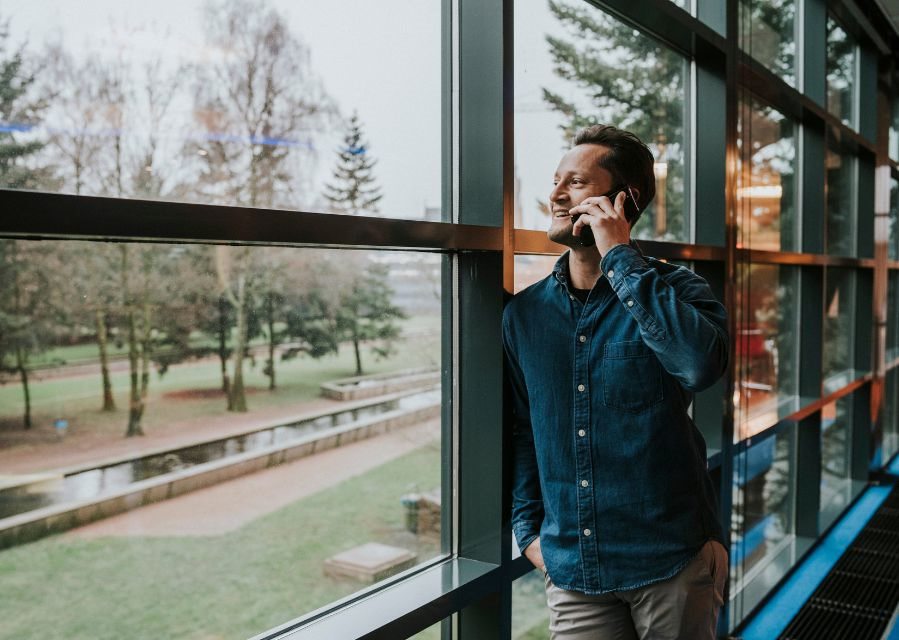 Een man leunt tegen een raamkozijn en is tegelijkertijd aan het bellen met een mobiele telefoon.