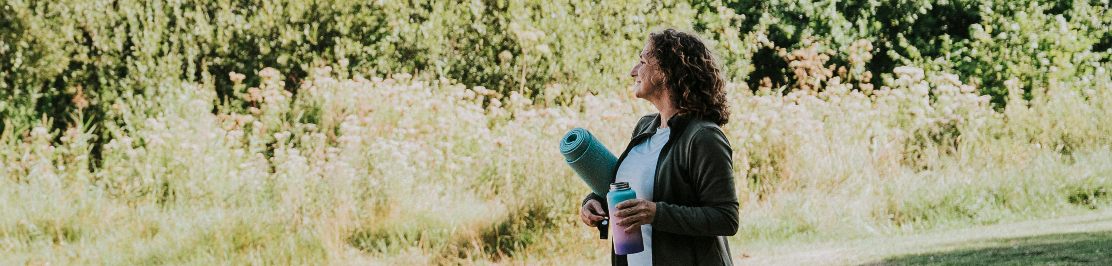 Vrouw loopt met een opgerolde yogamat onder haar arm en een drinkfles in haar hand door een park.