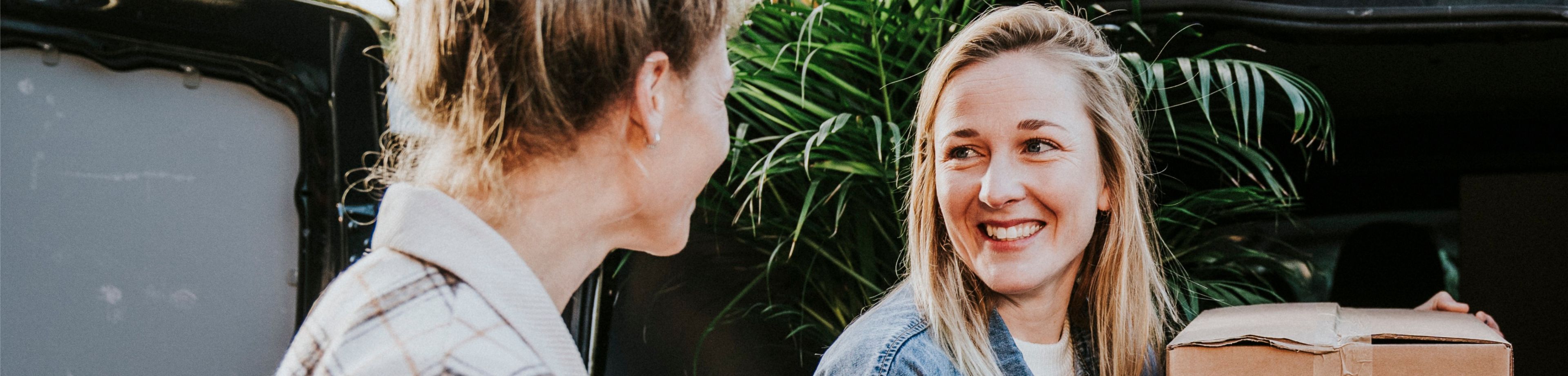 2 vrouwen lachen naar elkaar terwijl ze verhuisdozen uit een auto tillen. 